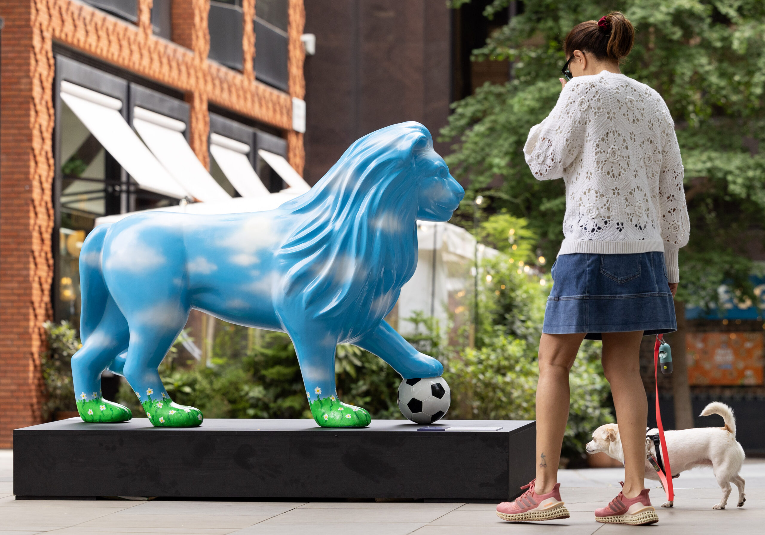 A woman in a white sweater and denim skirt walks a small dog past a colourful lion statue. The lion is painted with a sky pattern and stands on a black pedestal with a soccer ball at its foot. The background shows a brick building and green foliage, creating an urban oasis feel.