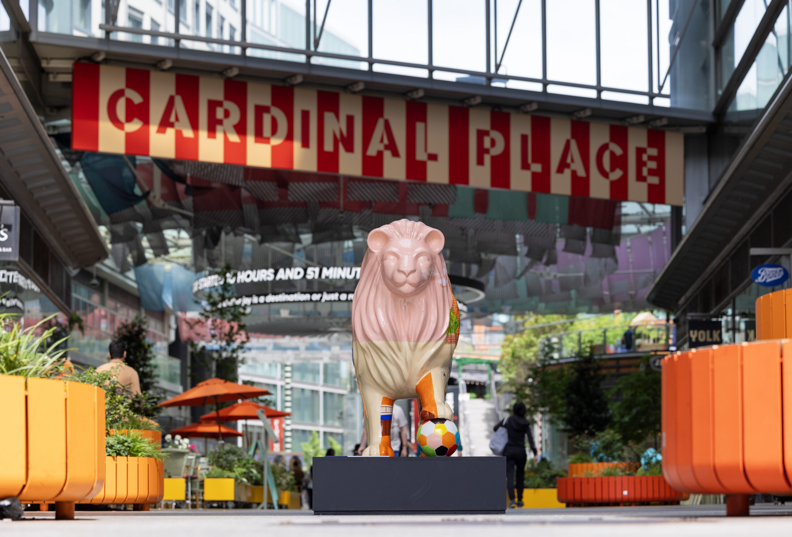 A colorful lion statue stands on a platform in a vibrant urban setting, with a "Cardinal Place" sign overhead. Bright orange planters and lively decorations surround it.