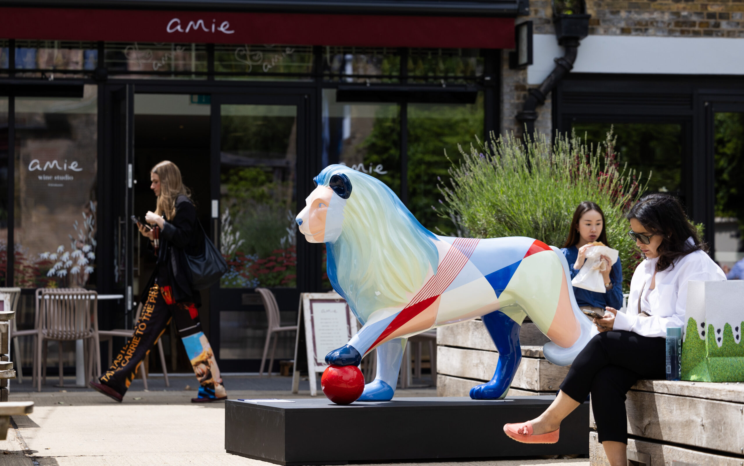 Colorful lion sculpture with geometric patterns stands in Eccleston Yards, with amie wine bar in the background. Two women sit on a bench nearby, one on a phone and the other eating, conveying a lively urban scene.