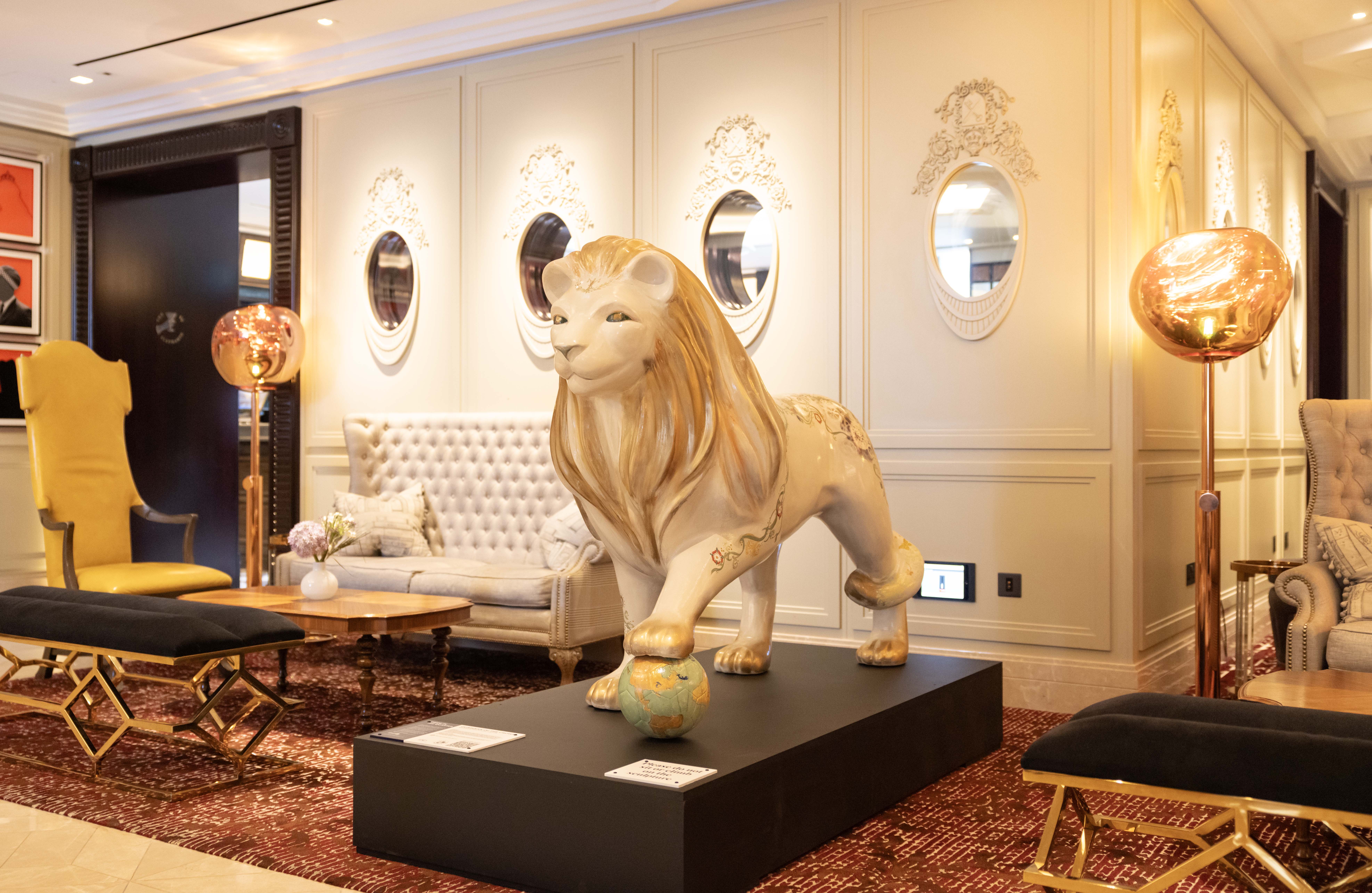 Elegant hotel lobby (Great Scotland Yard) with a lion statue on a pedestal, flanked by chic sofas and golden floor lamps. The atmosphere is luxurious and refined.