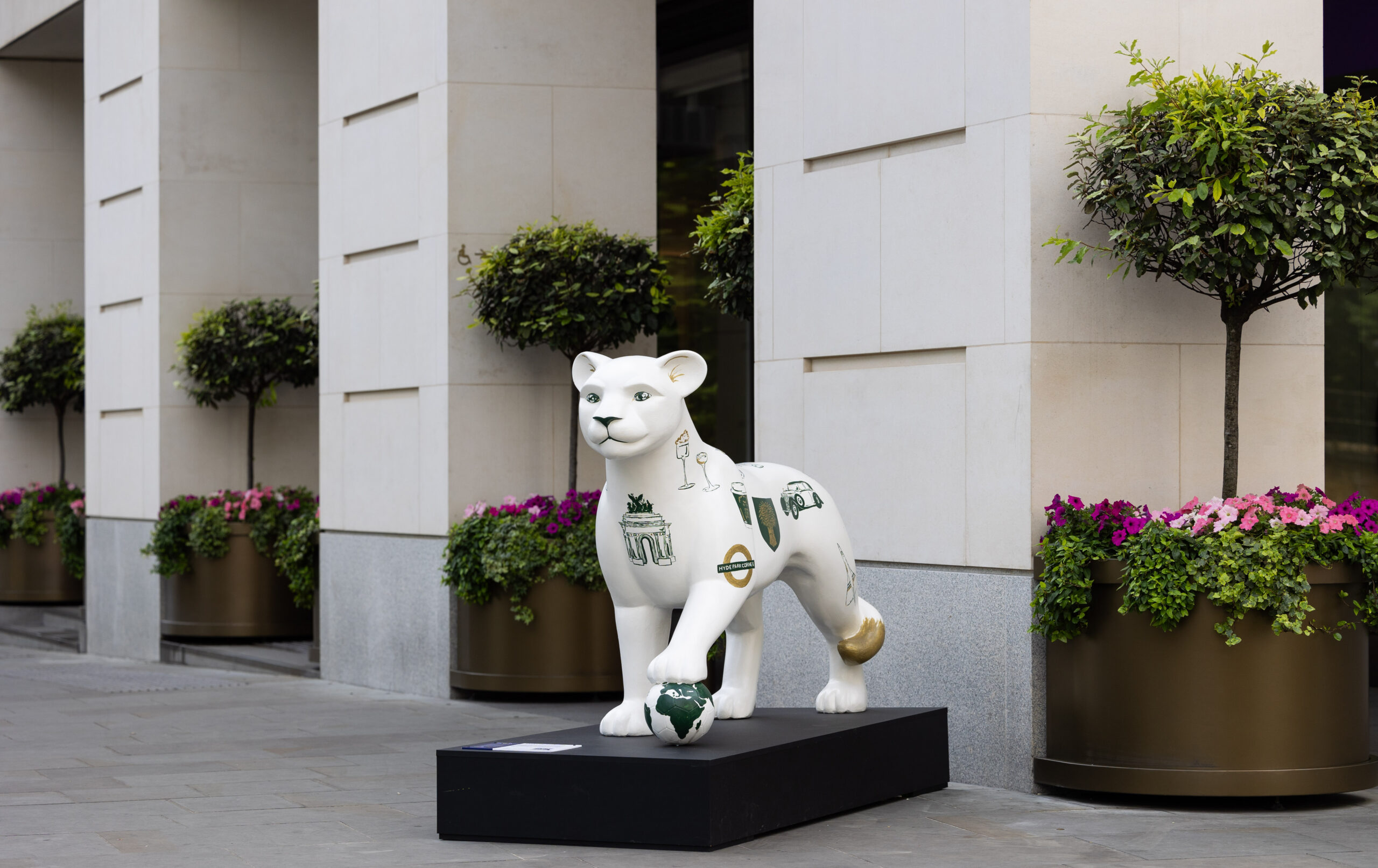 A white lion statue with decorative green symbols and a gold patch stands on a black pedestal. It's positioned in front of The Peninsula, a luxury hotel with lined potted plants.