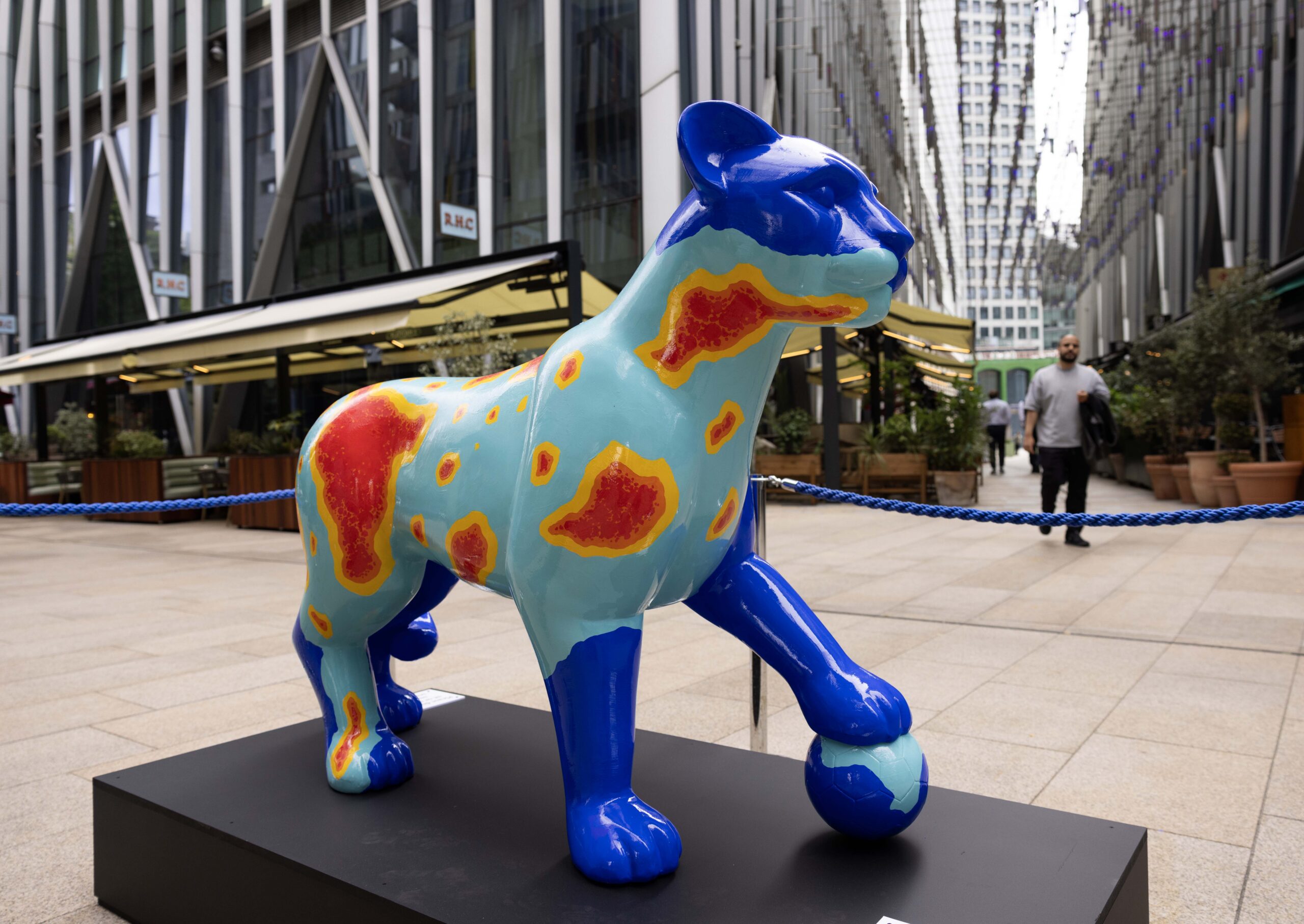 A vibrantly coloured leopard sculpture stands on a black platform in an urban courtyard, with a man walking by. The piece is bright blue with red and yellow spots, creating a striking visual against the modern building backdrop.