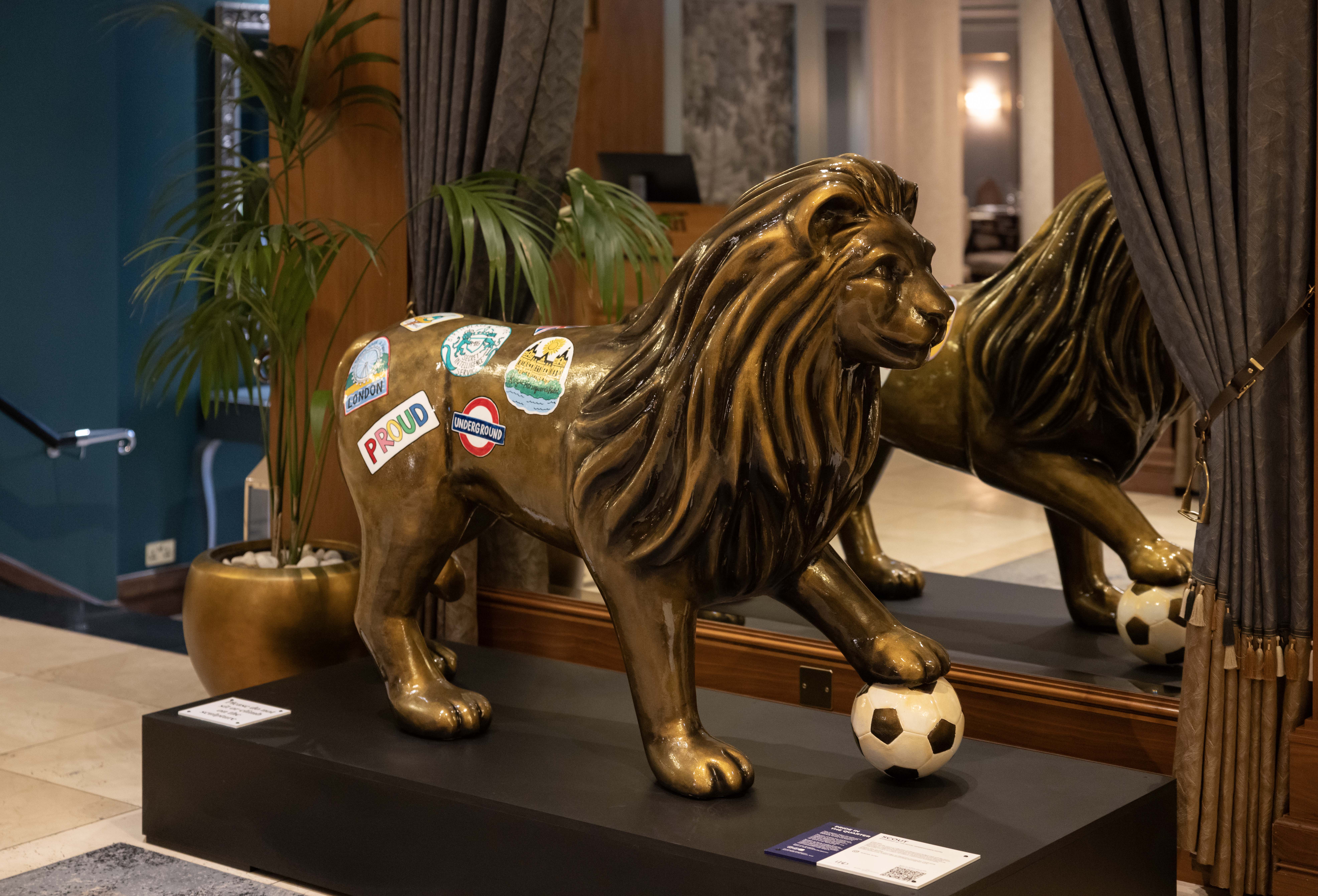 A golden lion statue with stickers stands on a black platform, its paw on a soccer ball. It is reflected in a mirror, surrounded by elegant décor in the Royal Horseguards Hotel.