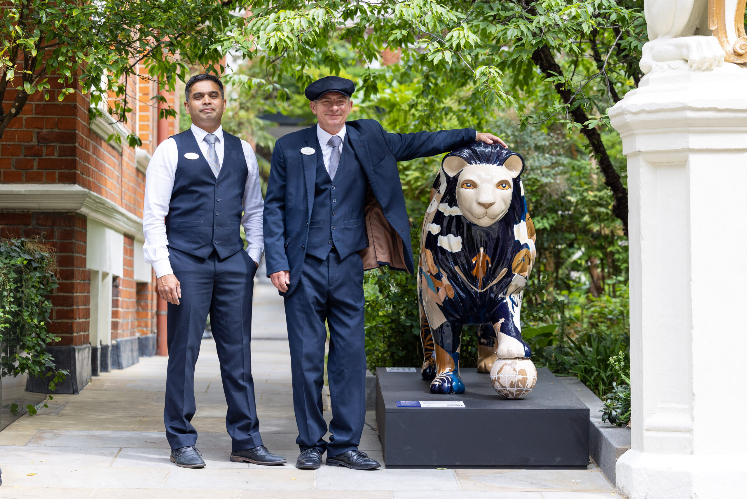 Two men in smart uniforms stand beside a colorful lion sculpture outdoors, surrounded by greenery and brick buildings, creating a formal yet friendly atmosphere.