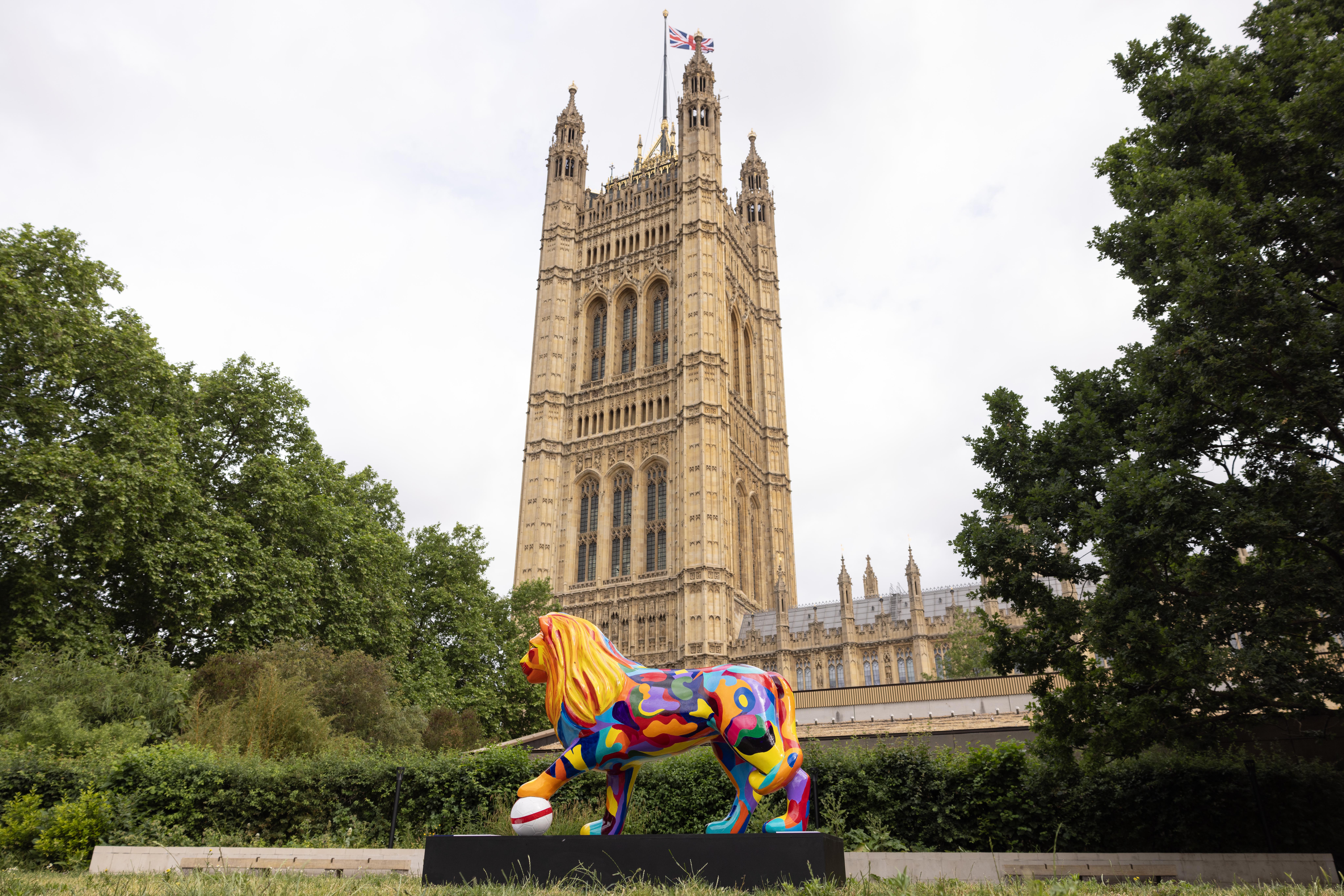 A vibrantly painted lion sculpture stands on a platform, with the majestic Gothic tower of the Palace of Westminster in the background, surrounded by greenery.