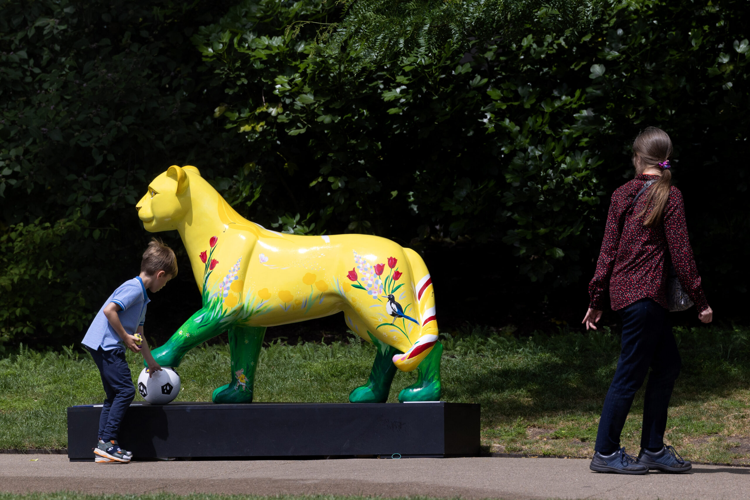 A young boy leans towards a colorful, floral-decorated yellow lioness statue holding a football. A girl walks nearby on a sunlit pathway.
