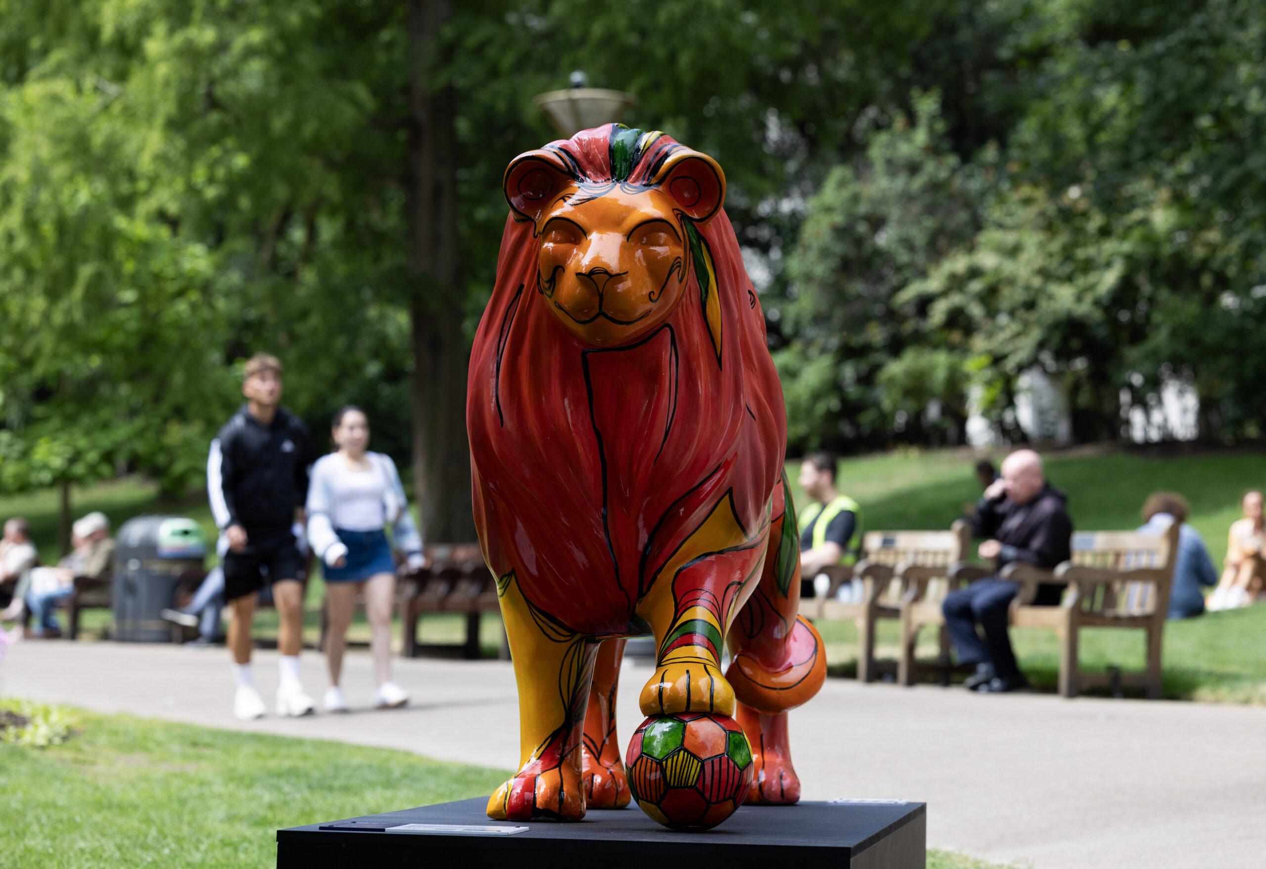 A colourful lion sculpture with a red mane and painted designs stands on a platform in a park. People stroll and sit on benches in the lush, green background.