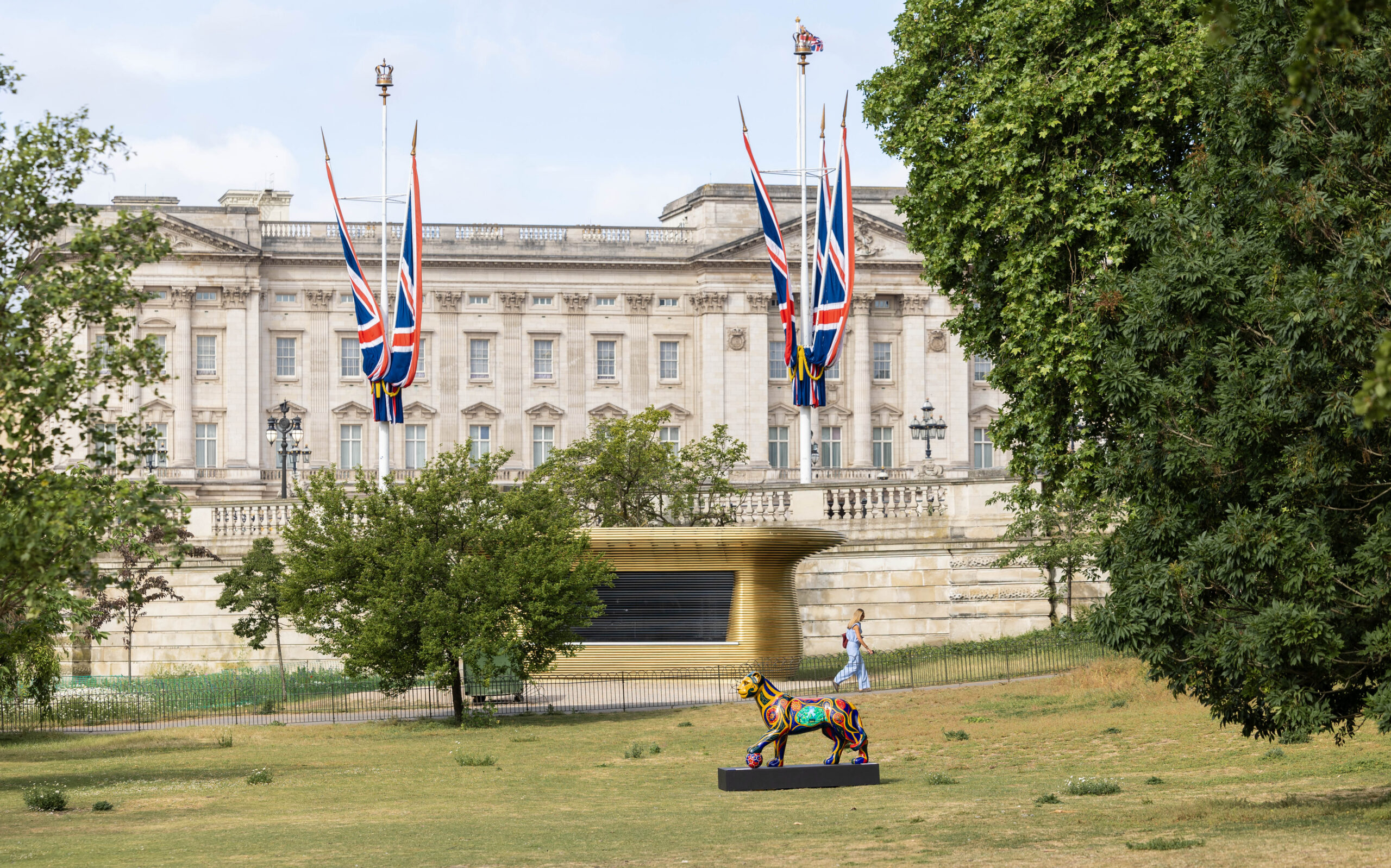 A colorful sculpture of a horse stands on a grassy area in front of Buckingham Palace adorned with Union Jack flags. Trees frame the scene, conveying a regal ambiance.